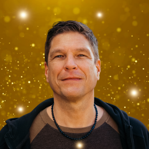 Ein Mann mit kurzen Haaren und einer Halskette steht vor einem goldenen Hintergrund mit kleinen, leichten Partikeln und erinnert an die inspirierende Atmosphäre beim TRAUMgeburt nach TRAUMAgeburt-Kongress zu modernen Geburtshilfetechniken.