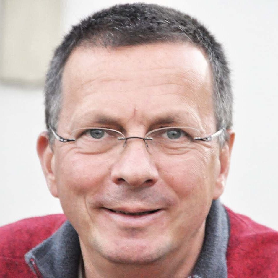 Ein Mann mit kurzen grauen Haaren, Brille und roter Jacke lächelt in die Kamera.