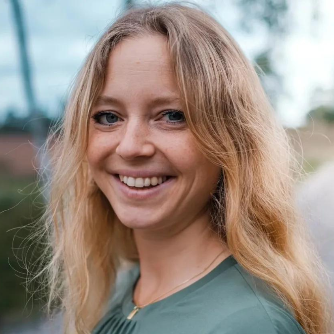 Eine Frau mit langen blonden Haaren und grünen Augen steht im Freien vor einem verschwommenen grün-blauen Hintergrund und lächelt in die Kamera. Sie trägt ein grünes Oberteil und verkörpert damit die Gelassenheit, die in der Geburtspsychologie oft thematisiert wird.